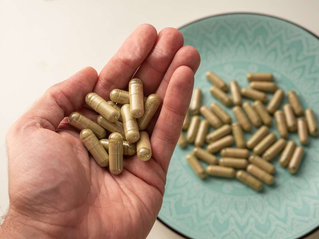 Mano che tiene capsule di kratom, con altre capsule su un piatto verde acqua